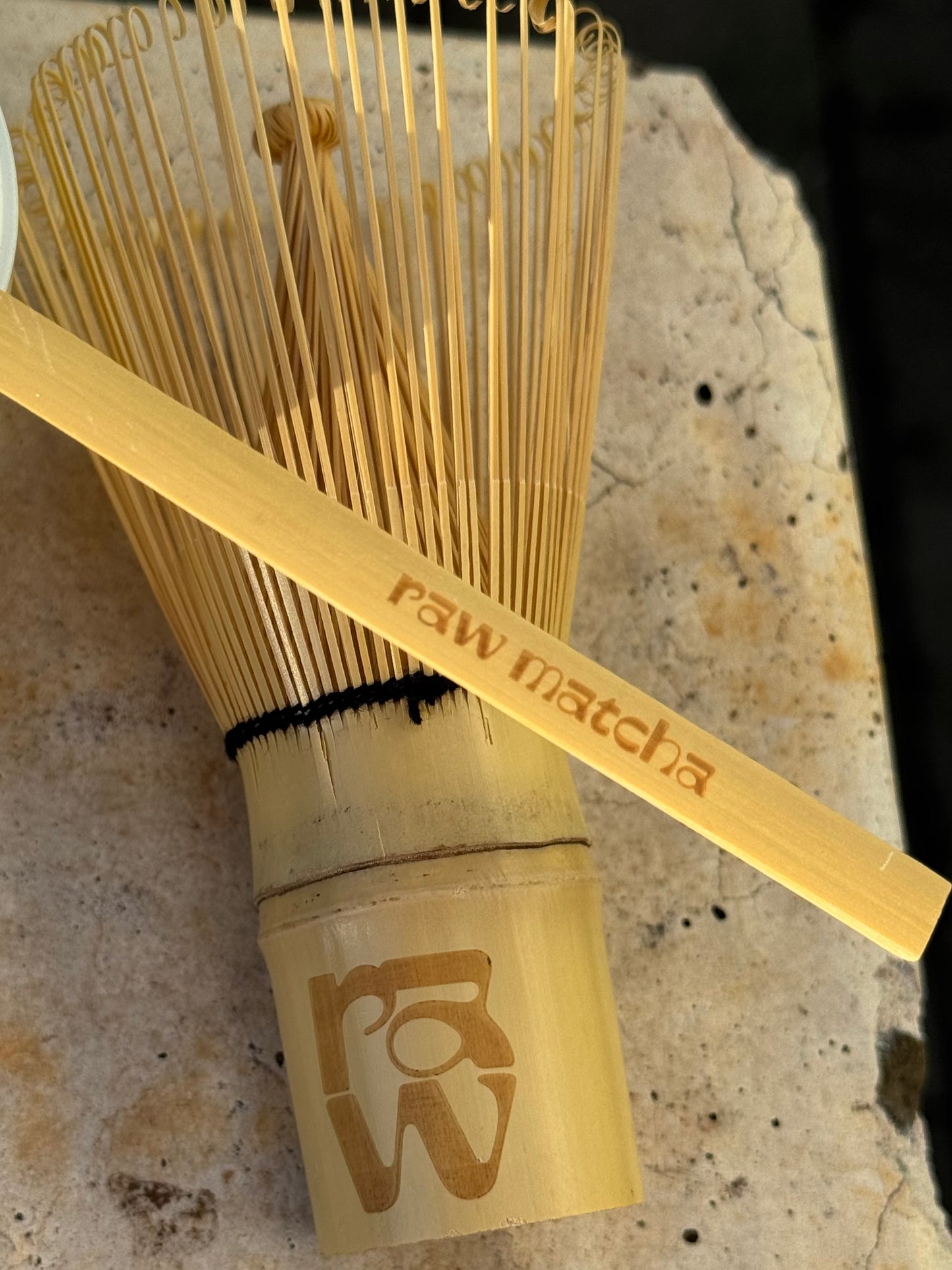 A bamboo match whisk and matching bamboo scoop resting on a textured stone surface. The bamboo scoop is engraved with the words 'raw matcha' and features a stylized 'RW' logo on the whisk's handle.