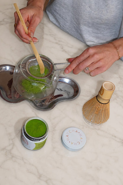 A person's hands are shown whisking green matcha tea in a glass teapot with a bamboo whisk and a small jar of matcha powder on a white marble countertop.